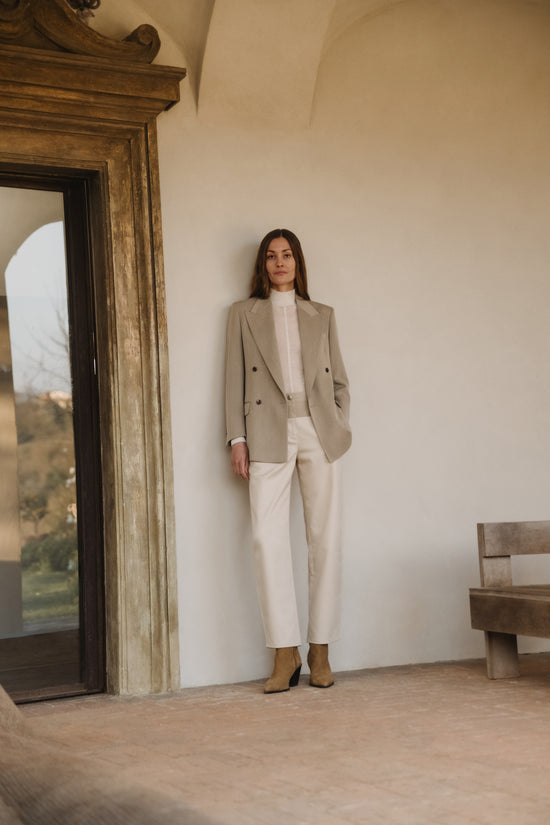 Woman leaning against a cream wall wearing a beige double-breasted blazer, a white high-neck top, ivory trousers and tan suede ankle boots