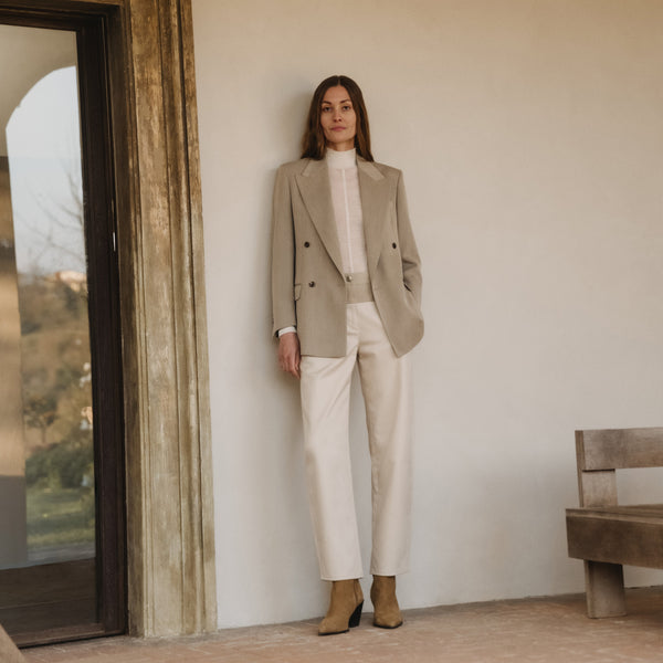 Woman leaning against a cream wall wearing a beige double-breasted blazer, a white high-neck top, ivory trousers and tan suede ankle boots