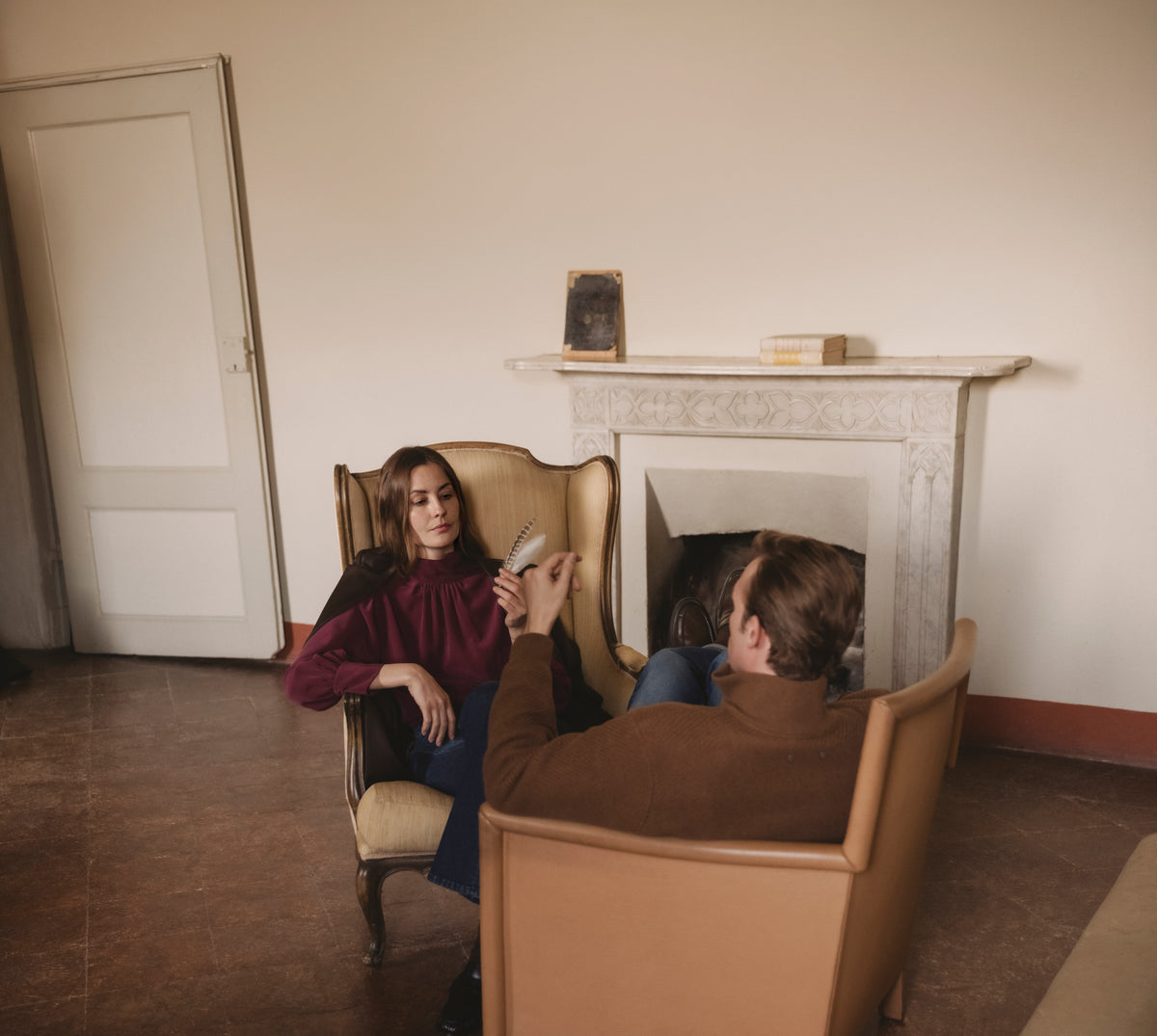 Woman in a burgundy high-neck blouse and dark jeans seated in an armchair, facing a man in a brown knit sweater and blue jeans by a marble fireplace