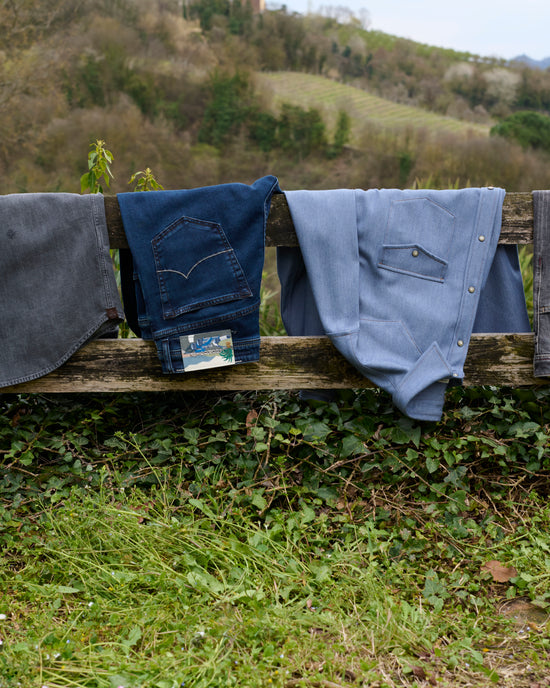 A pair of denim jeans in dark blue and two denim shirts:  light blue wash and grey, draped over a wooden fence with a countryside hillside backdrop