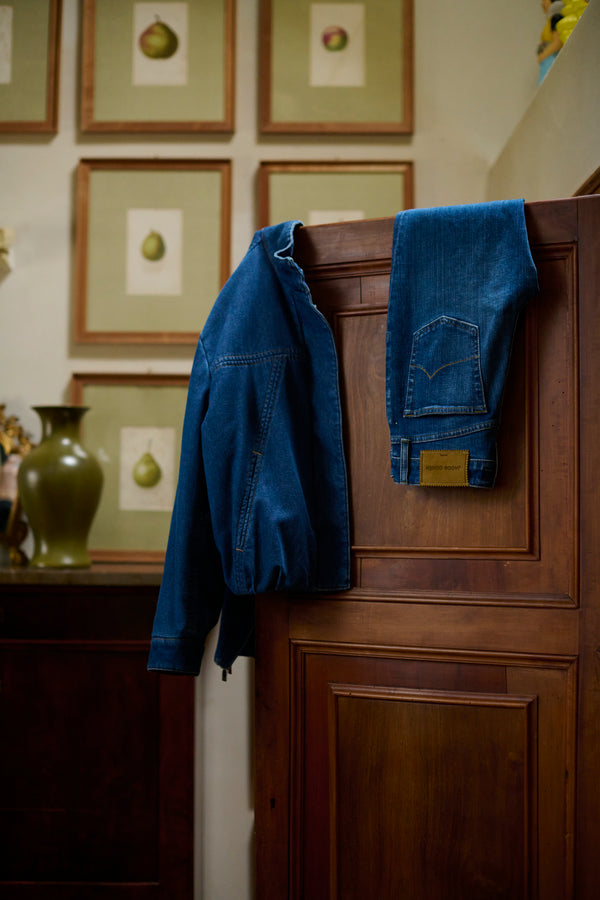 Denim jacket and jeans with leather patch hanging on a wooden cabinet, with framed botanical fruit prints and a ceramic vase in the background