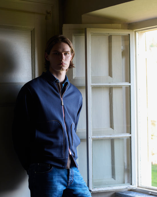 Model wearing a navy zip-up jacket with patch pockets and a striped contrast zip, styled with a blue collared denim shirt and denim jeans, standing by a white-shuttered window.