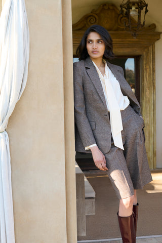 Model wearing a brown houndstooth-check blazer and matching Bermuda shorts, styled with a cream pussy-bow blouse and tall burgundy leather boots, posed in an arched doorway