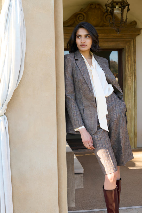 Model wearing a brown houndstooth-check blazer and matching Bermuda shorts, styled with a cream pussy-bow blouse and tall burgundy leather boots, posed in an arched doorway.
