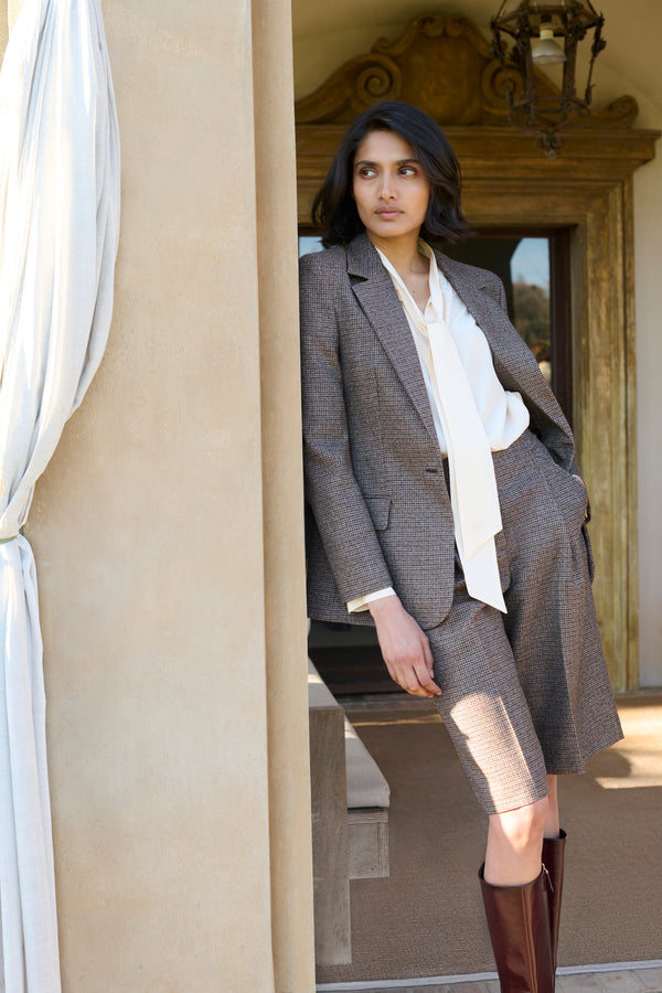 Model wearing a brown houndstooth-check blazer and matching Bermuda shorts, styled with a cream pussy-bow blouse and tall burgundy leather boots, posed in an arched doorway