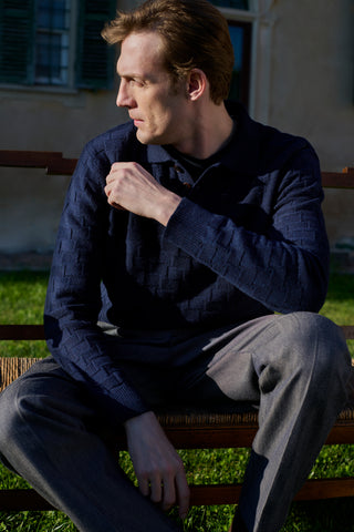 Man wearing a navy textured knit polo sweater and grey trousers, sitting on a wooden bench outdoors