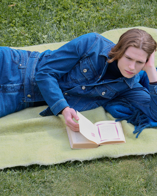 Model reclining on a lime-green fringed blanket in a total denim look, with a mid-wash denim jacket and matching jeans over a dark roll-neck, reading a book on the grass.