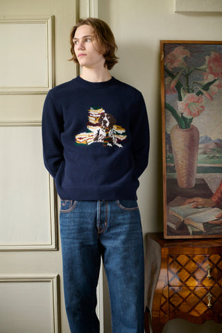 Man wearing a navy sweater with an intarsia dog and books motif, paired with denim jeans, standing indoors beside a vintage painting and antique wooden chest of drawers