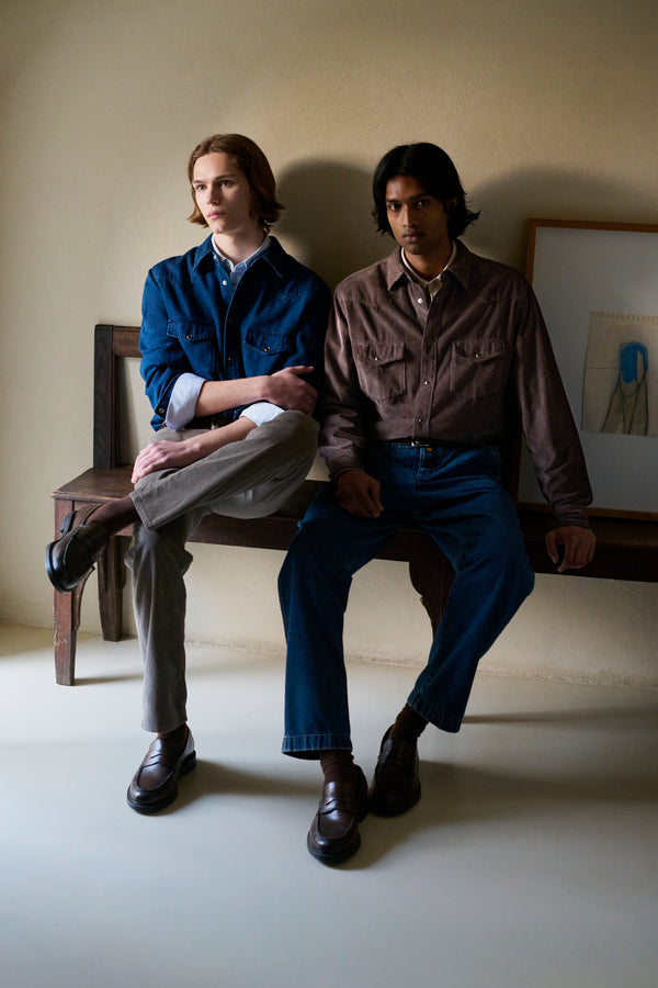 Two men seated on a wooden bench against a beige wall, one wearing a navy denim shirt with grey trousers, the other in a brown corduroy shirt with dark blue jeans, both in brown leather loafers, with a framed artwork in the background