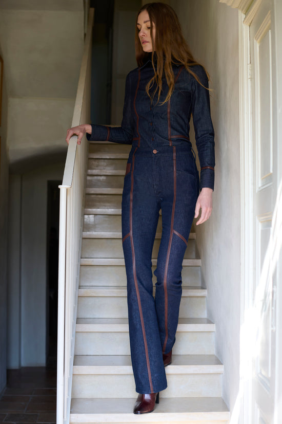 Woman on a staircase wearing a dark denim shirt and jeans with contrast leather piping seams, paired with burgundy leather boots, in a bright indoor stairwell setting.