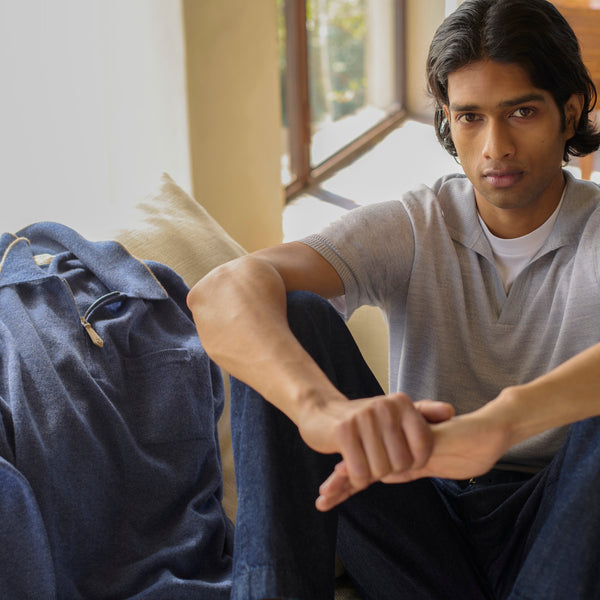 Man in a grey knit polo and dark denim seated by a window, hands clasped, with a navy zip-up jacket draped beside him on the sofa