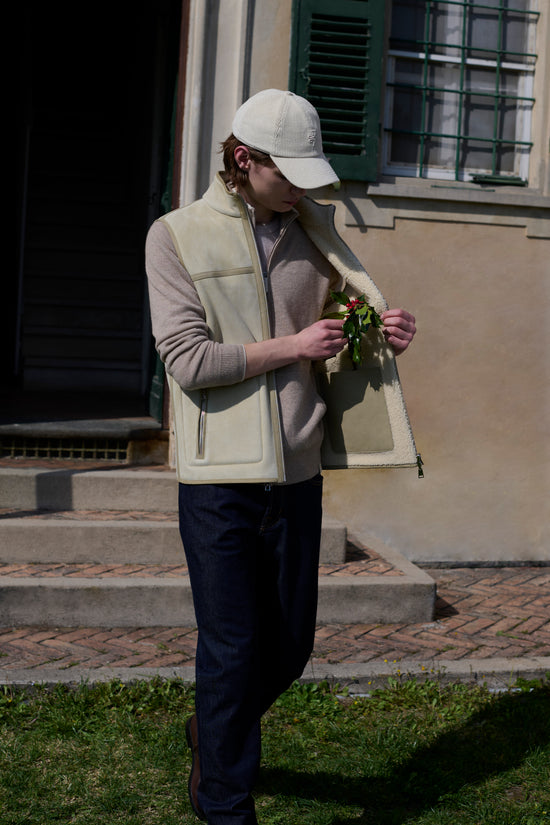 Man in a cream corduroy cap, a cream shearling vest, a beige knit sweater and dark jeans standing outdoors holding a sprig of holly