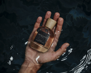 Hand emerging from dark water holding a glass perfume bottle with a cork cap and dark textured label