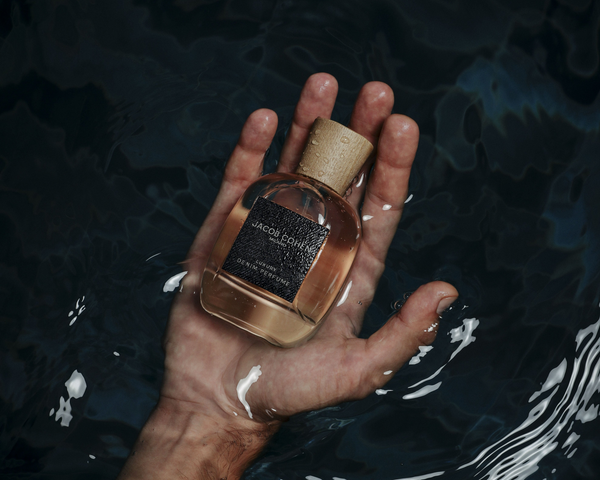 Hand emerging from dark water holding a glass perfume bottle with a cork cap and dark textured label