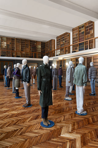 Historic double height library converted into a fashion showroom: a spacious hall with light-and-dark Hungarian herringbone parquet flooring and white mannequins displaying a elegant autumn collection in neutral tones, browns, grays, navy blue, and forest green