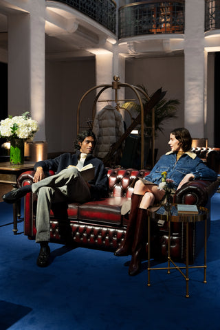 Five star hotel lobby with double height classical architecture: two guests in smart casual attire converse on a burgundy Chesterfield sofa