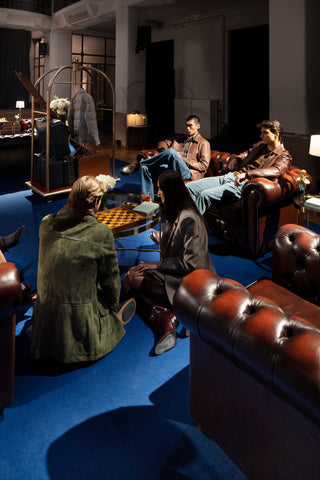 Group of four people conversing in a five star hotel lounge: two seated on a leather sofa (a man in a brown jacket with jeans, a man in a brown jacket with light blue trousers) and two in the foreground seen from behind (a woman with dark hair, a man in an olive-green jacket)