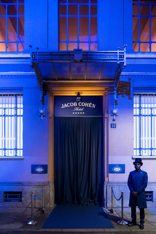 Main entrance of a luxury hotel at night: a white facade with tall windows illuminated from inside with warm light, bathed in blue exterior lighting