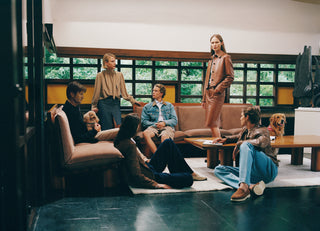 Group of six people in contemporary casual wear relaxing in modern living room with beige sofas, wooden coffee table, and two dogs, overlooking garden through large windows