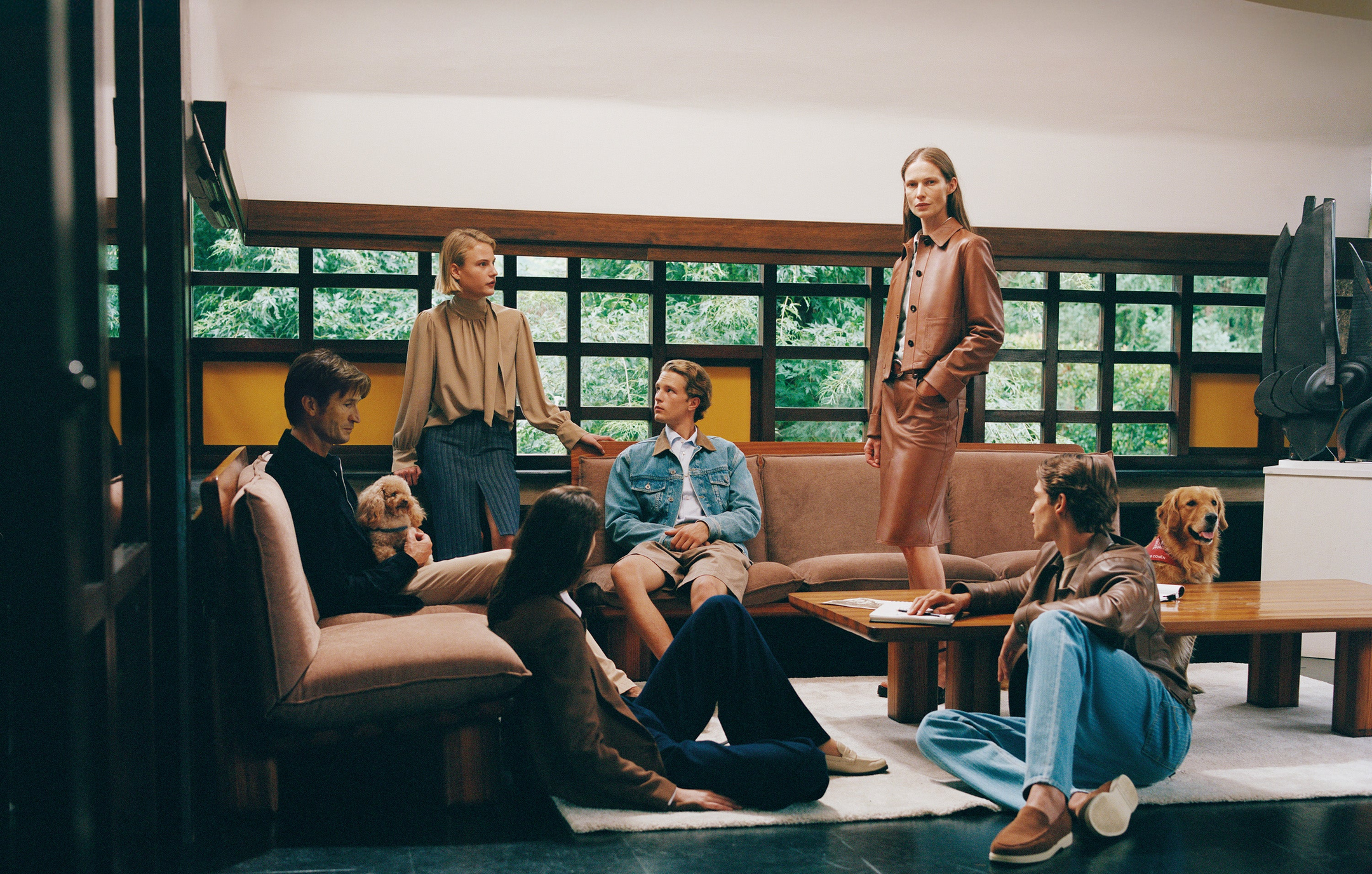 Group of six people in contemporary casual wear relaxing in modern living room with beige sofas, wooden coffee table, and two dogs, overlooking garden through large windows