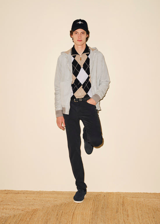 A young man wearing a blue and white argyle sweater paired with a light blue jacket, blue pants, and black shoes. He's also wearing a black baseball cap with a white logo. The background is a plain beige wall with a wooden floor.