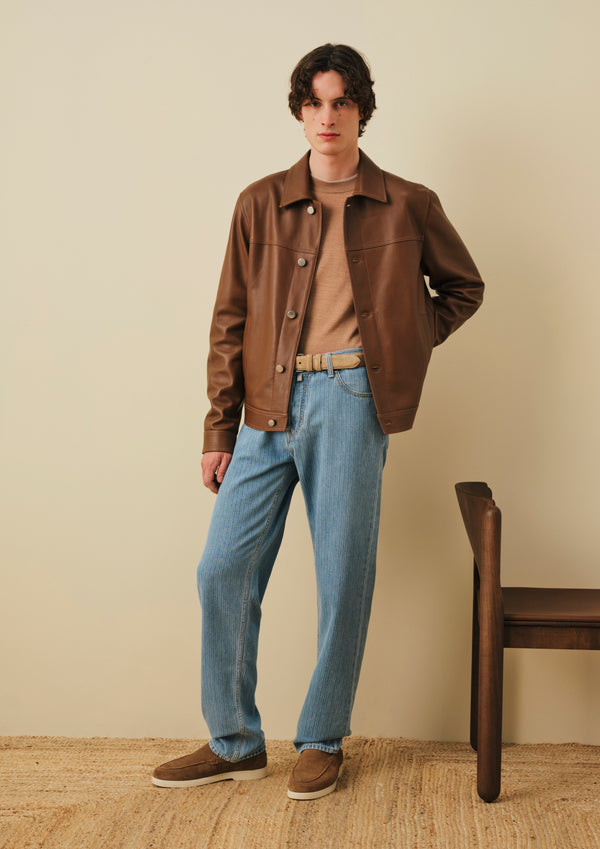 Model wearing a brown work jacket over a beige sweater, light blue relaxed-fit jeans with a tan belt, and brown suede sneakers, standing against a neutral beige background