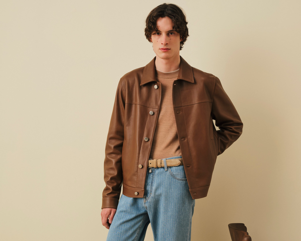 "Young man with dark wavy hair wearing a brown canvas jacket over a tan turtleneck sweater, light blue corduroy pants, and a beige belt, standing against a beige background in a fashion photoshoot pose