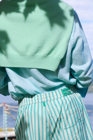 Close-up back view of a Jacob Cohën outfit featuring a mint green knit sweater layered over a light blue shirt, paired with green and white striped swimwear, shot against a coastal backdrop