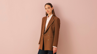 Woman wearing camel brown double-breasted blazer over white shirt with blue wide-leg trousers against pink background