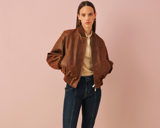Young woman with long straight dark hair and gold hoop earrings wearing a brown satin bomber jacket over a cream silk blouse and dark blue jeans, standing against a soft pink background in a fashion photoshoot pose