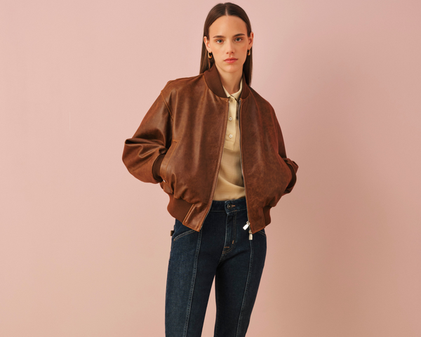 Young woman with long straight dark hair and gold hoop earrings wearing a brown satin bomber jacket over a cream silk blouse and dark blue jeans, standing against a soft pink background in a fashion photoshoot pose