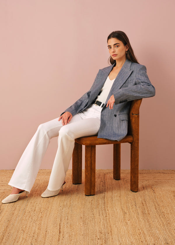 A young woman sitting on a wooden chair against a soft pink wall, wearing a blue double-breasted blazer over a white top with a black belt, paired with white wide-leg pants