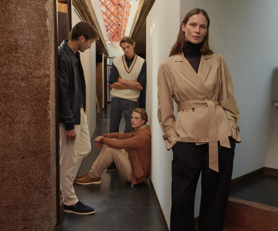  Four models showcasing autumn wardrobe essentials: beige wrapped blazer with belt, brown cardigan, cream knit vest, and blue bomber jacket, styled with neutral trousers in a contemporary setting