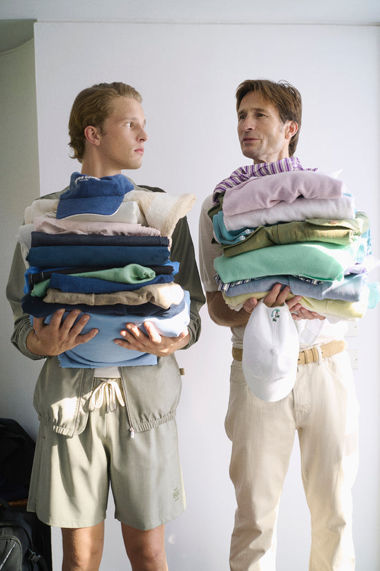 Two male models holding large stacks of folded knitwear and casual garments in pastel and earthy tones. One wears sage green drawstring shorts, the other cream linen trousers with a canvas belt. Playful editorial setting with a white background.