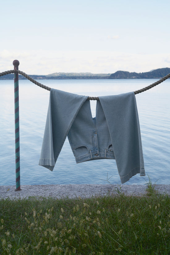Light wash denim jeans hanging to dry over a rope by a calm lake, with hills in the background and grass in the foreground.