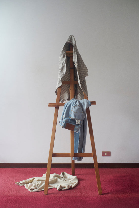 Wooden easel used as a clothes rack displaying a grey checkered oversized shirt draped over the top, light blue denim jeans hanging in the middle, and a cream knit garment casually dropped on the red carpet below