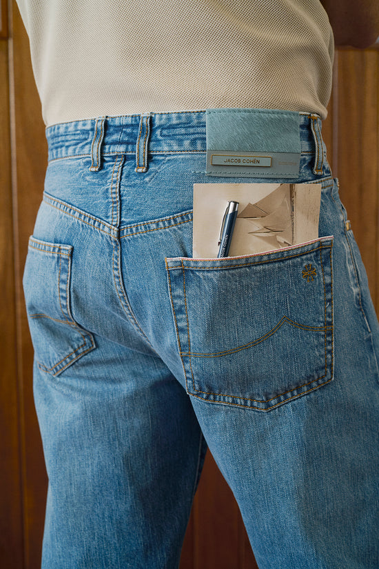 Close-up back view of light wash denim jeans with a Jacob Cohën logo patch, featuring a back pocket with a pen and folded papers tucked inside, paired with a cream knit top.