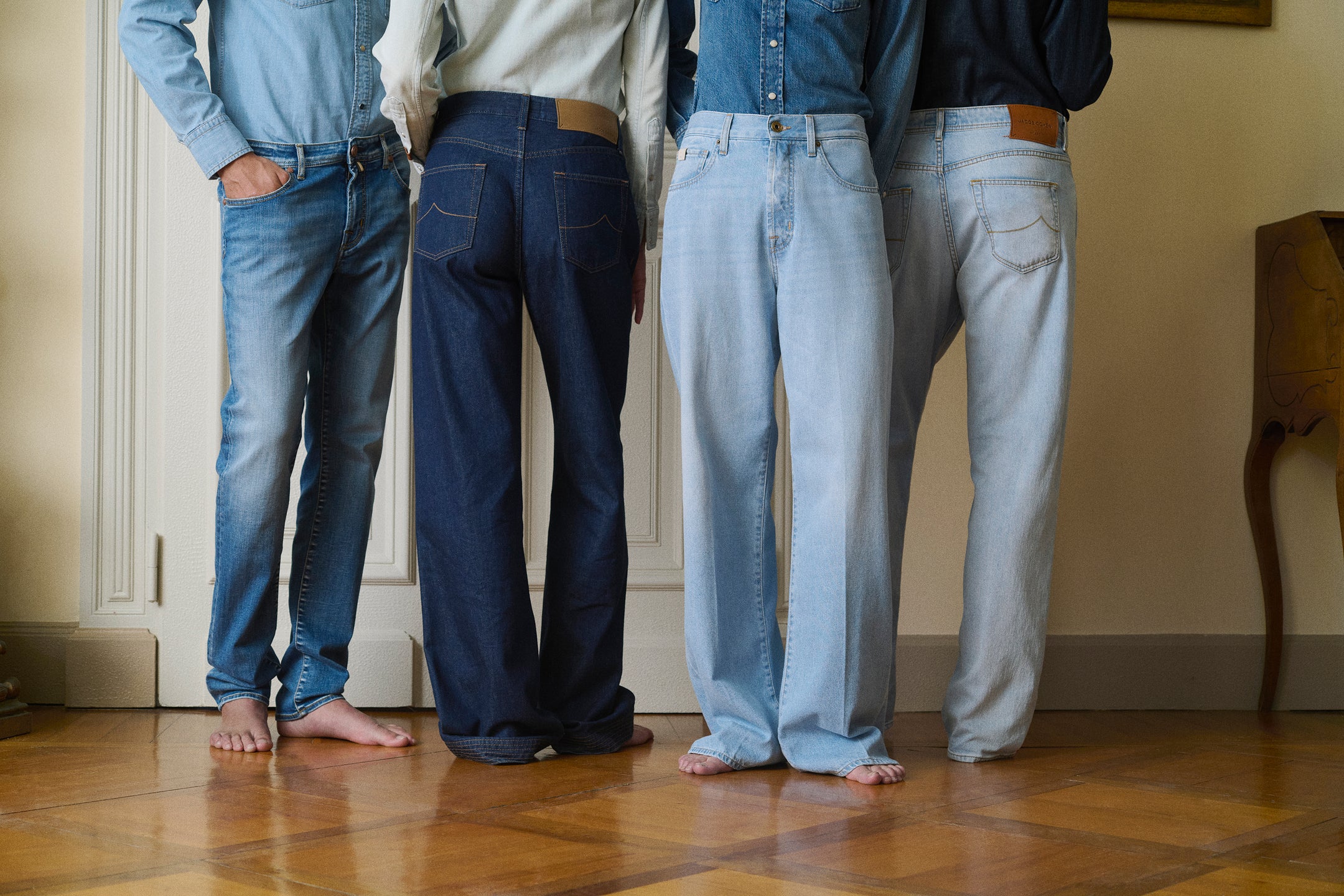 Four models standing side by side, shot from behind, each wearing a different style of denim jeans ranging from dark indigo to light wash, paired with denim shirts, barefoot on a parquet floor
