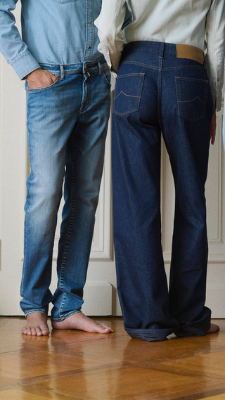 Two models standing side by side, shot from behind, wearing different denim jeans styles: a slim-fit mid-wash blue jean on the left and a wide-leg dark indigo jean with contrast stitching and a tan leather patch on the right, both barefoot on a parquet floor