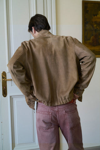 Rear view of a man wearing a tan suede bomber jacket and washed pink jeans, standing in front of a white door