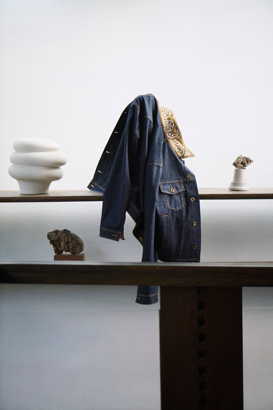 Dark indigo denim jacket with ornate gold embroidery detail on the collar, draped over a wooden shelf alongside decorative ceramic and stone objects