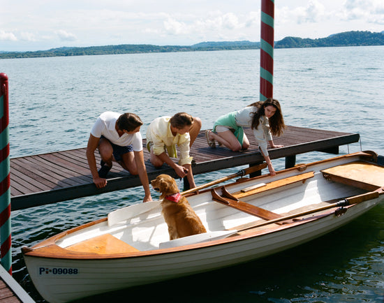 Three models dressed in summer casual outfits — white t-shirt with navy shorts, yellow shirt with bermuda shorts, and a light top with light shorts — gathered on a lakeside dock around a wooden rowing boat with a golden retriever. Outdoor lifestyle setting.