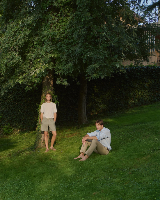 Two male models in a garden setting — one standing by a tree wearing a cream short-sleeve knit top and olive drawstring shorts, the other sitting on the grass in a light blue linen shirt and taupe trousers
