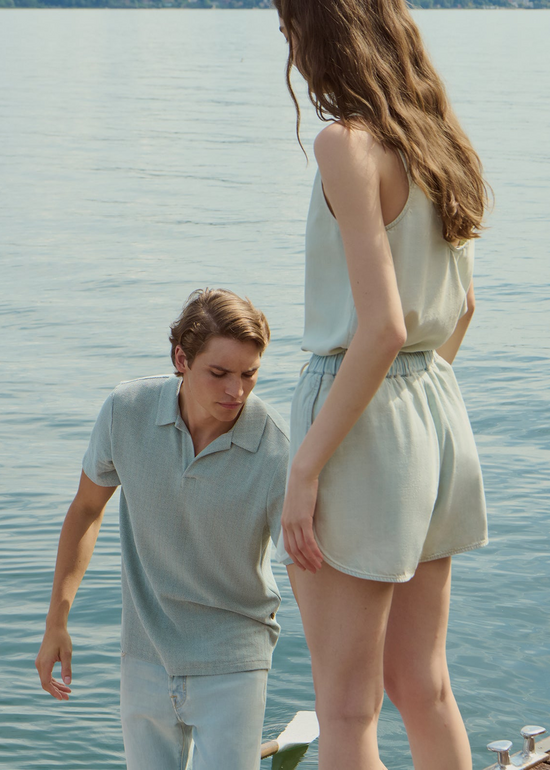 A young man and woman standing on a dock by calm water. He wears a light grey textured polo shirt with pale blue jeans, she wears a light blue denim top and matching shorts.
