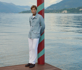 Man leaning against a red and green striped nautical pole on a wooden dock, wearing a light blue linen overshirt with patch pockets over a white t-shirt, white trousers and dark loafers