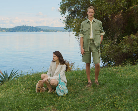 Man and woman posing outdoors by a lake. The woman is crouching on the grass, wearing a cream blazer and shorts, holding a light blue mesh bag. The man is standing, wearing a green overshirt jacket with matching shorts and brown loafers. Natural setting with trees and water in the background.