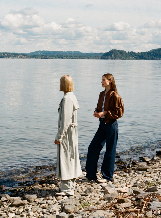 Two people standing on a lakeshore wearing contemporary, minimalist outfits featuring a long light trench coat and a brown leather jacket paired with wide-leg trousers.