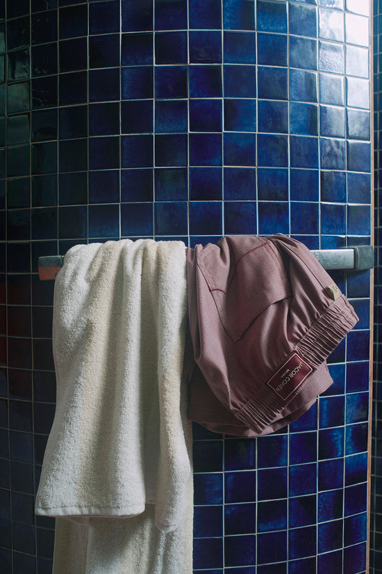 Mauve drawstring swimshorts hanging on a rail against a blue tiled wall.