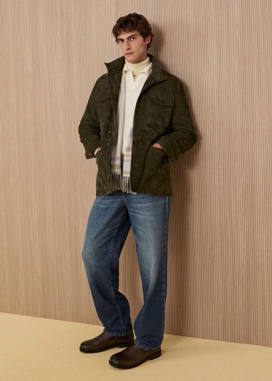 Model leaning against a wall wearing an olive green suede field jacket, a cream polo shirt with a striped scarf, wide-leg medium-wash jeans, and ankle boots.