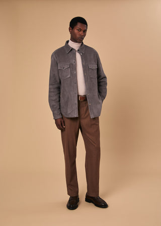 Man wearing a grey suede shirt jacket over a striped turtleneck, brown trousers, and dark loafers, standing against a beige backdrop
